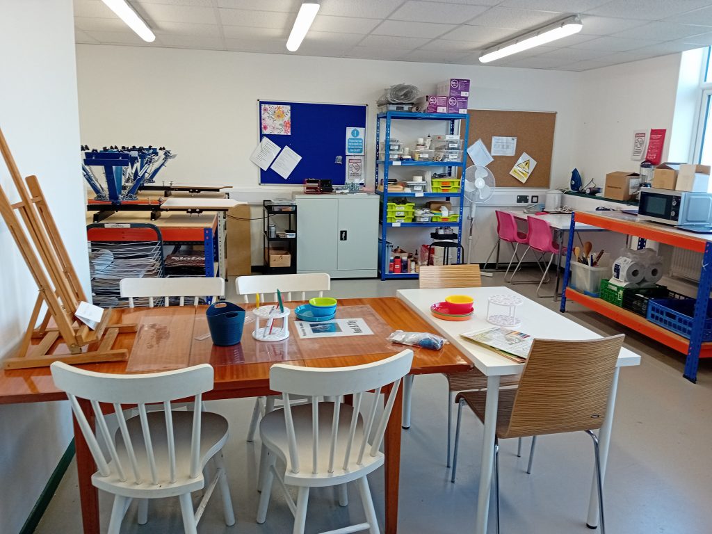 The image shows a craft room with a large table work area, screen printing stations and a Batik wax station.